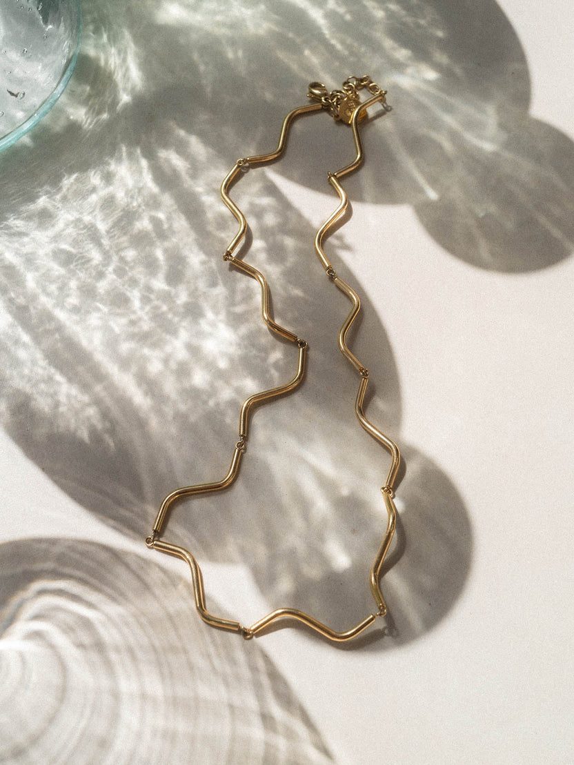 Wellenförmige, goldene Kette von PURELEI auf hellem Hintergrund mit sanften Schatten.