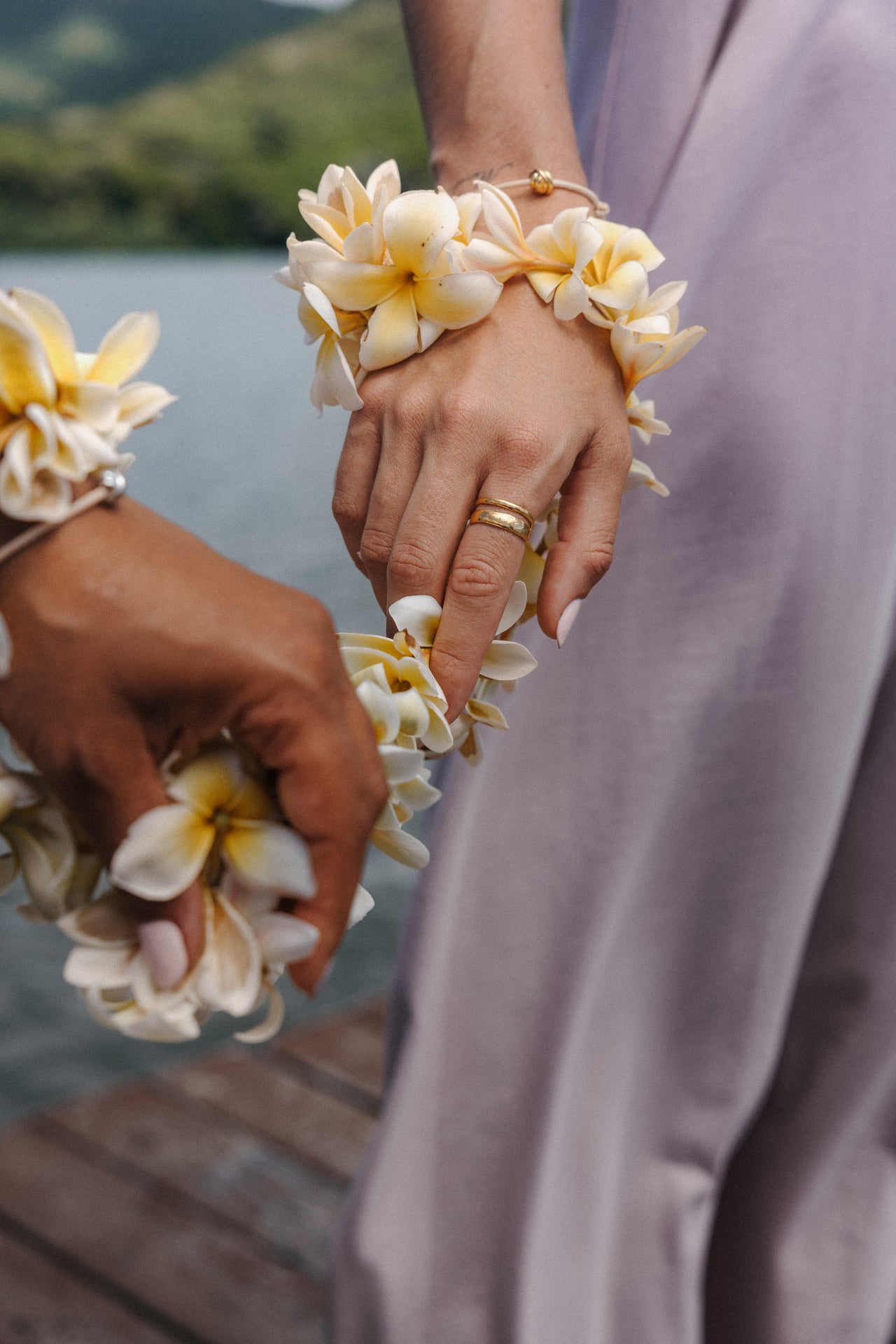 Zarte PURELEI Armbänder aus weißen Blüten, getragen von zwei Händen, umgeben von tropischer Stimmung.