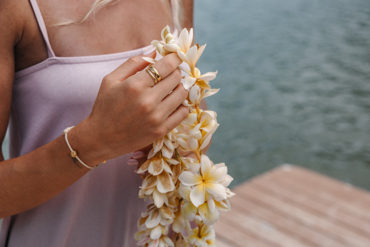 Goldring und Armband von PURELEI, getragen mit einer Blütenkette am Wasser.