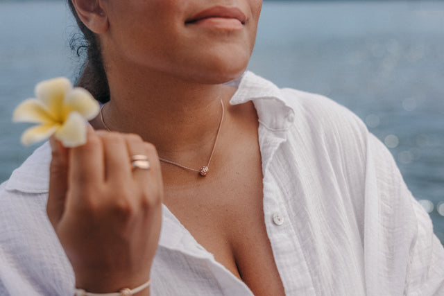 Roségoldene PURELEI Kette mit Kugel-Anhänger, getragen mit Blüte in der Hand, am Wasser.