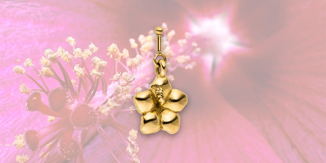 Goldener Blumenanhänger von PURELEI vor zartem pinken Hintergrund.