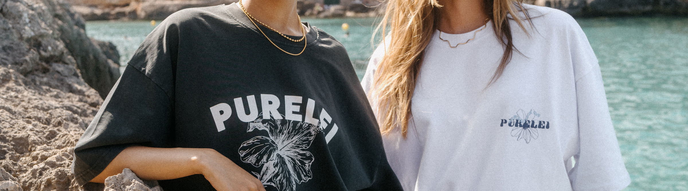 Zwei Frauen in PURELEI T-Shirts, eines schwarz mit Blumenmotiv, das andere weiß mit Logo, am Strand.