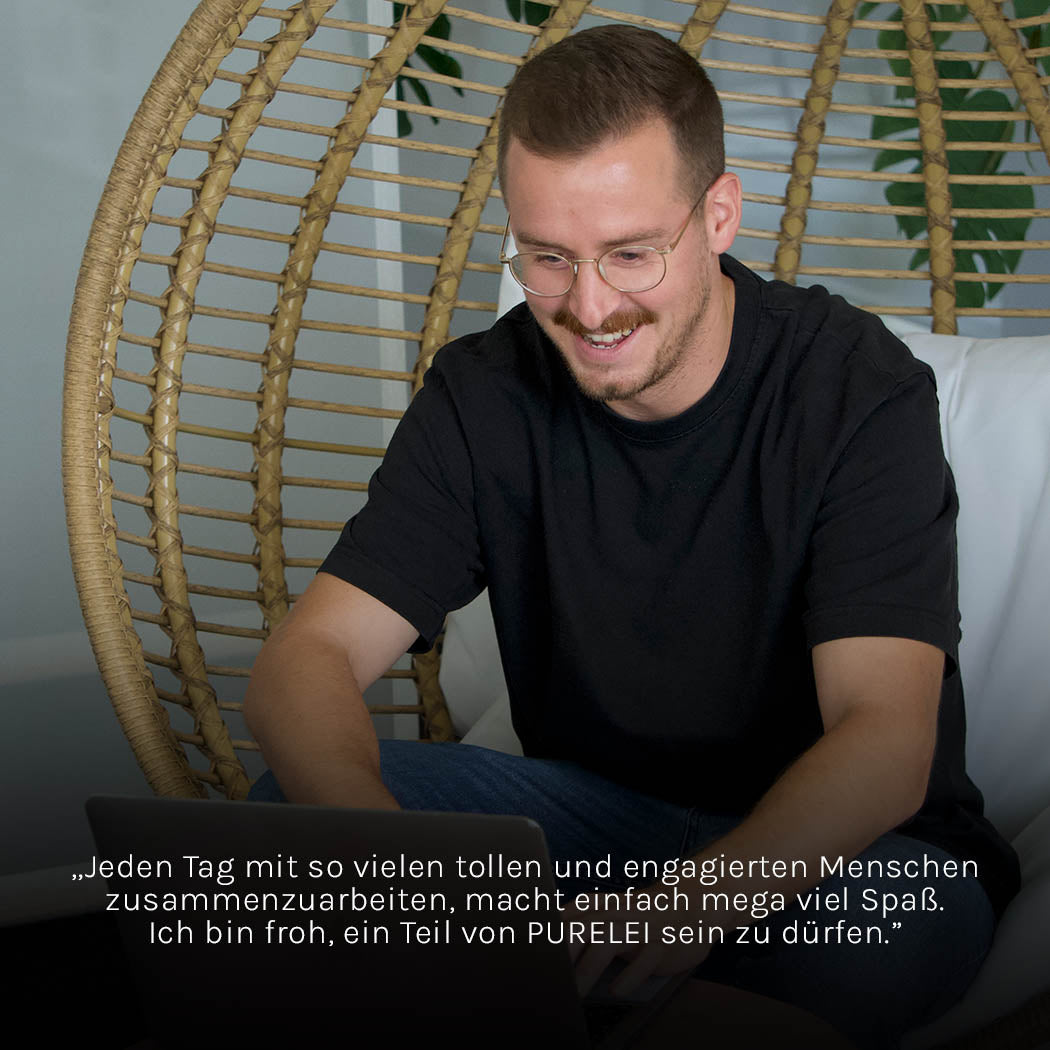 Mann in schwarzem Shirt lächelt, Teil von PURELEI, entspannt in einem geflochtenen Stuhl.