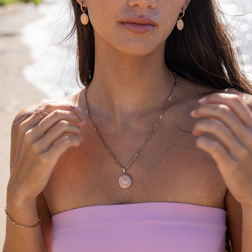 PURELEI Waina Ohrringe aus rosévergoldetem Edelstahl mit filigranem Charm, elegant am Strand getragen.