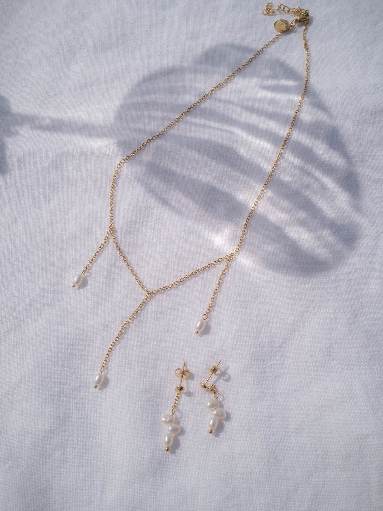 PURELEI Mahina Club Purely Y-Kette und Ohrringe in 18K Gold mit natürlichen Süßwasserperlen auf weißem Stoff