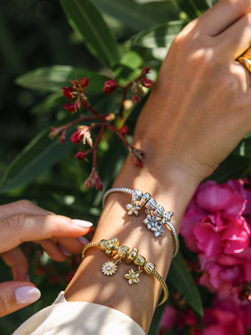 PURELEI Lei Plumeria Charm in Silber und Gold mit abnehmbarer Blüte, sichtbar am Handgelenk vor Blumenkulisse