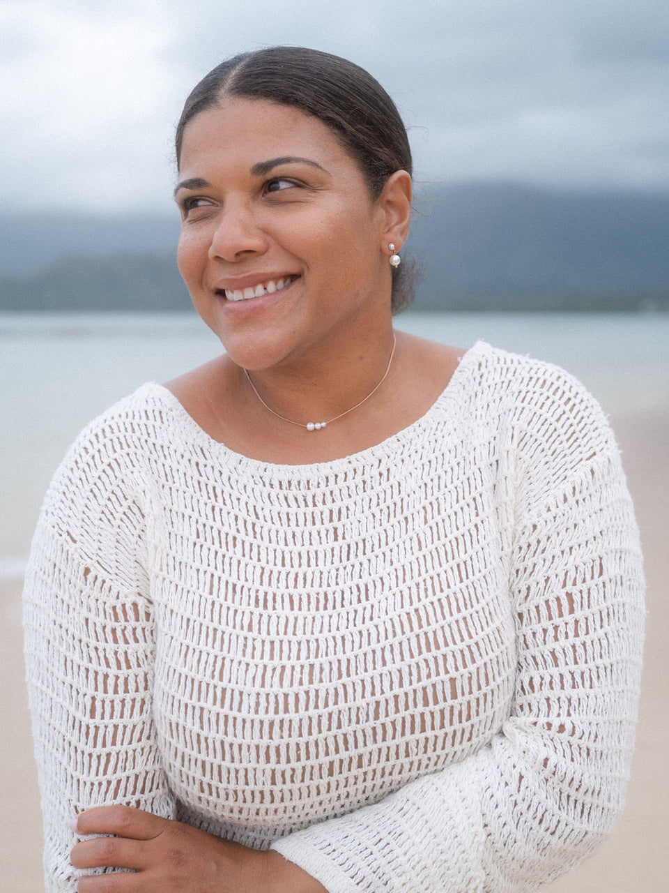 PURELEI Perlenkette und Ohrringe, elegant und feminin, getragen am Strand mit sanften Wellen im Hintergrund.