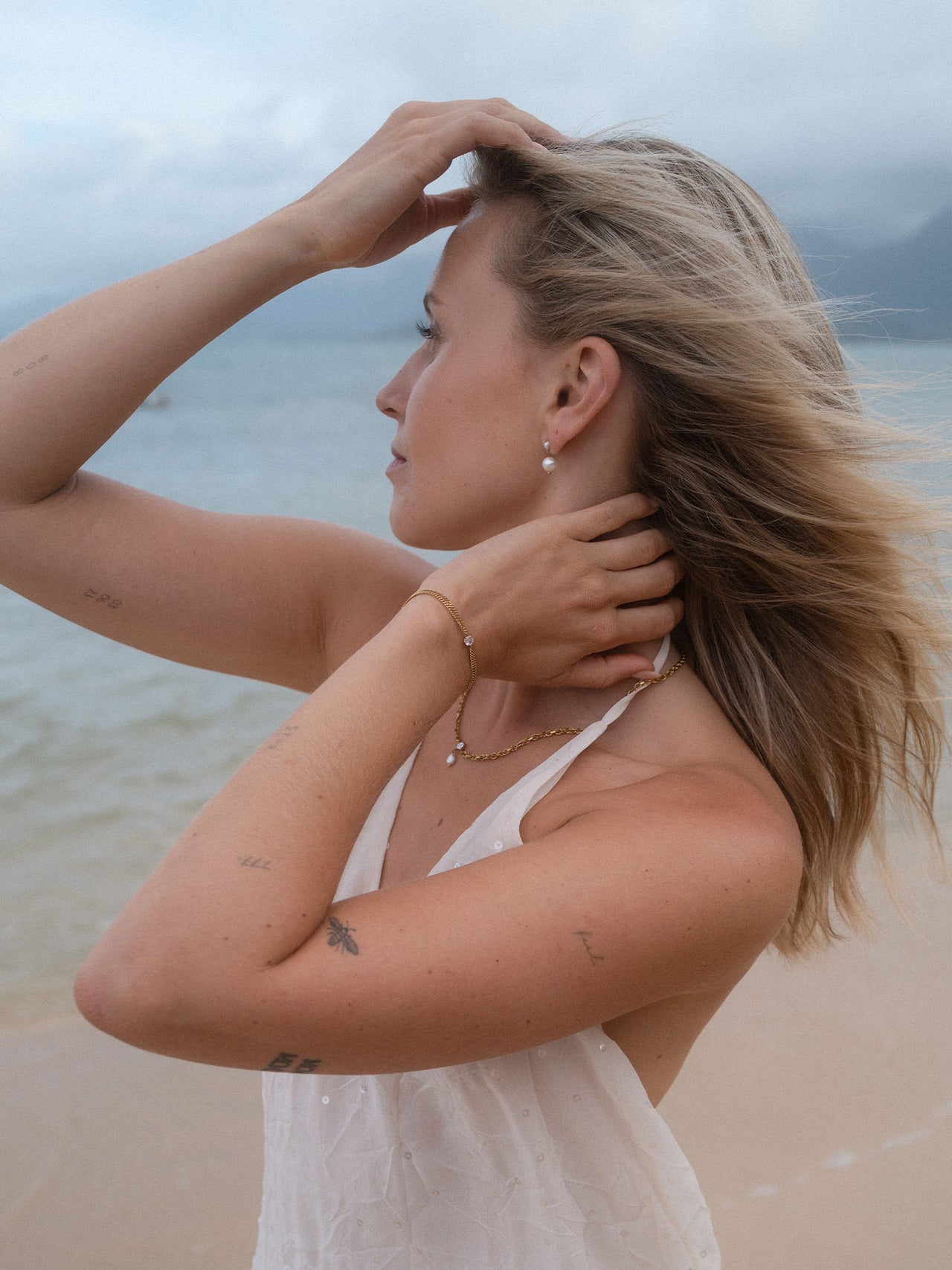 Elegante PURELEI Perlenohrringe und zartes Armband am Strand, umgeben von sanften Wellen.