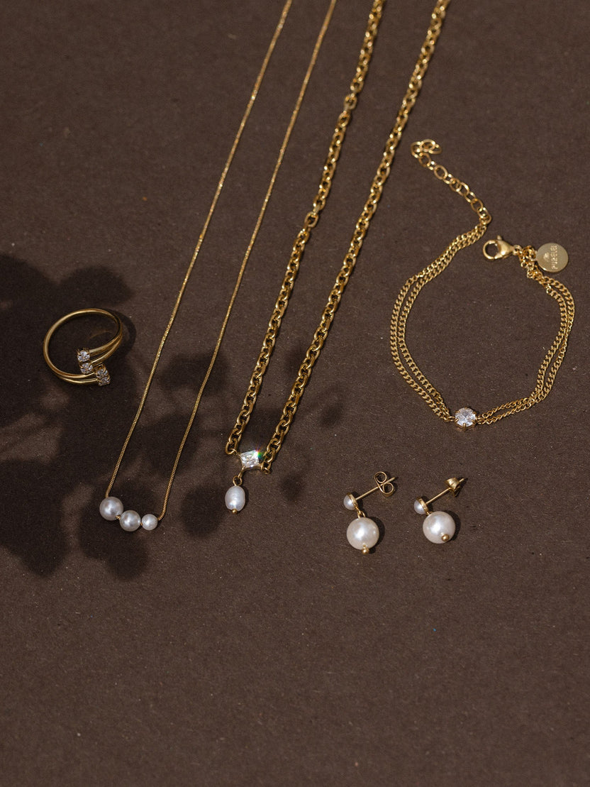 Goldene PURELEI Schmuckstücke: Kette, Armband, Ring und Ohrringe auf dunklem Hintergrund.